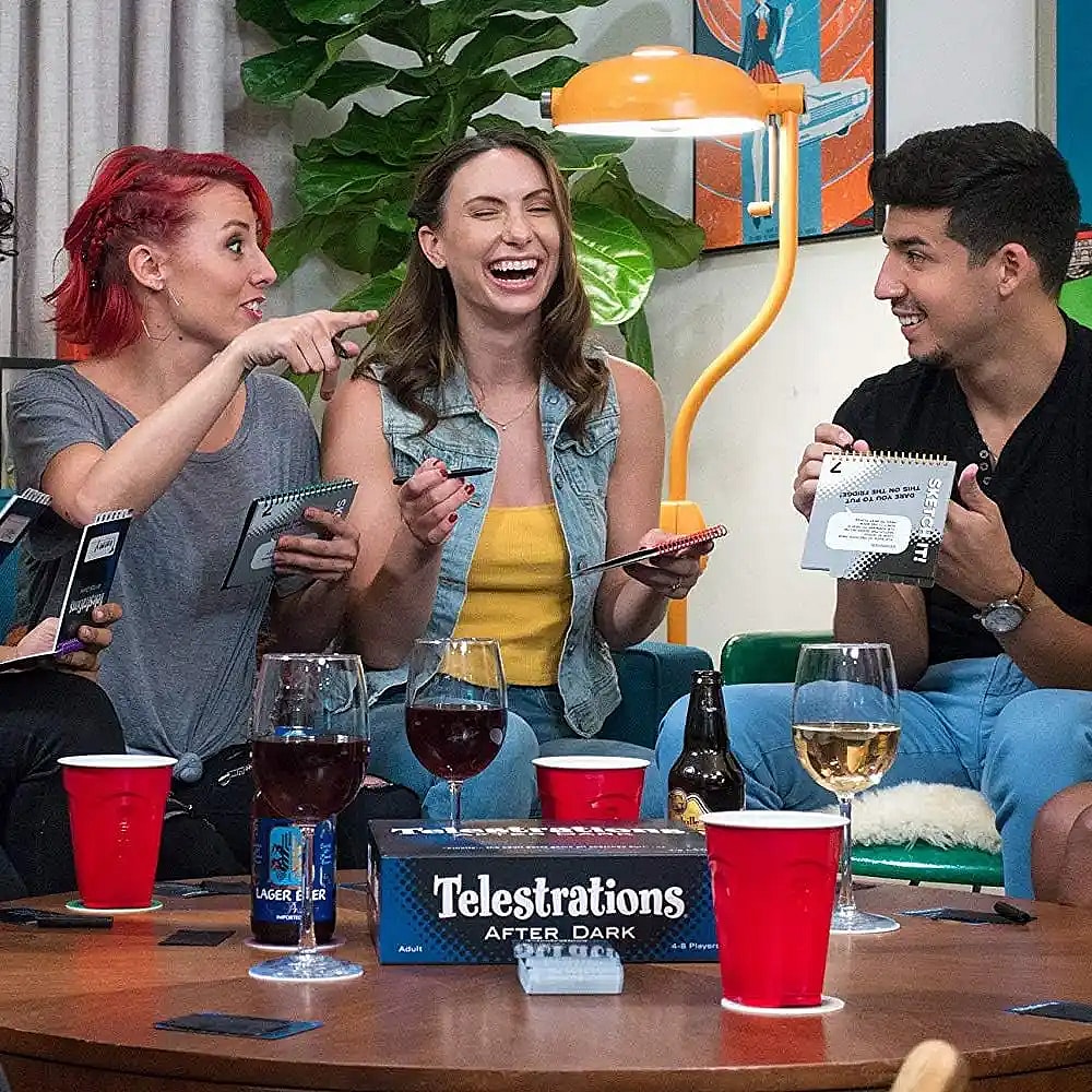 A group of four adults is sitting around a round wooden table, laughing while playing the board game Telestrations After Dark. Three of the people are holding open spiral-bound notebooks and pens. The woman with red hair on the left is pointing and smiling at the woman in the middle, who is laughing with her head tilted back. The man on the right is also smiling while holding his book and pen. On the table are several red plastic cups, a bottle of beer, a can of beer, and glasses of red and white wine. The game box is visible on the table in the center.
