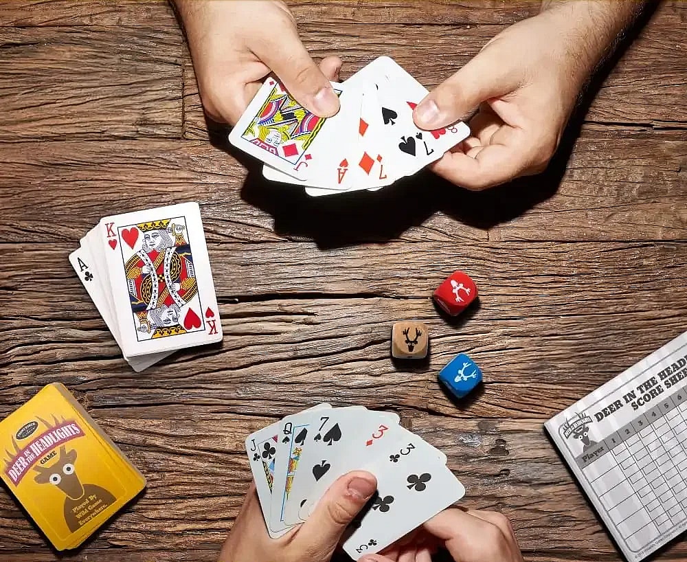 Overhead view of 2 people playing Deer in the Headlights card and dice game on a wooden table. The game is setup with the draw pile, 3 dice in the center, and the discard pile visible.