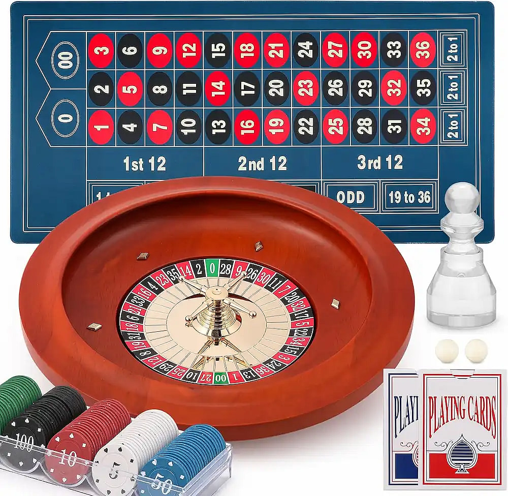 A high-angle shot shows a wooden roulette set with a felt betting mat featuring a double-zero American layout. The set includes a professional-style wheel, stacks of colorful poker chips in a clear tray, two decks of playing cards, two white roulette balls, and a clear plastic win marker.