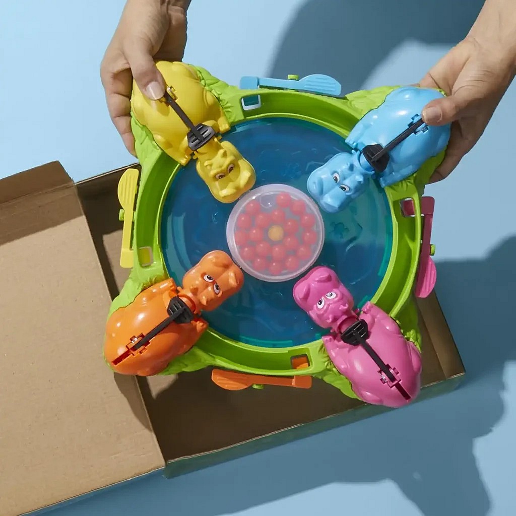 A pair of hands is holding the Hungry Hungry Hippos game above an open cardboard box, showing the four colorful plastic hippos (yellow, blue, orange, and pink) positioned around the central pool of marbles on a blue background. The second image is the box cover for the Aeon's End (Second Edition) board game, which features a group of mages battling a massive, red, horned demon.