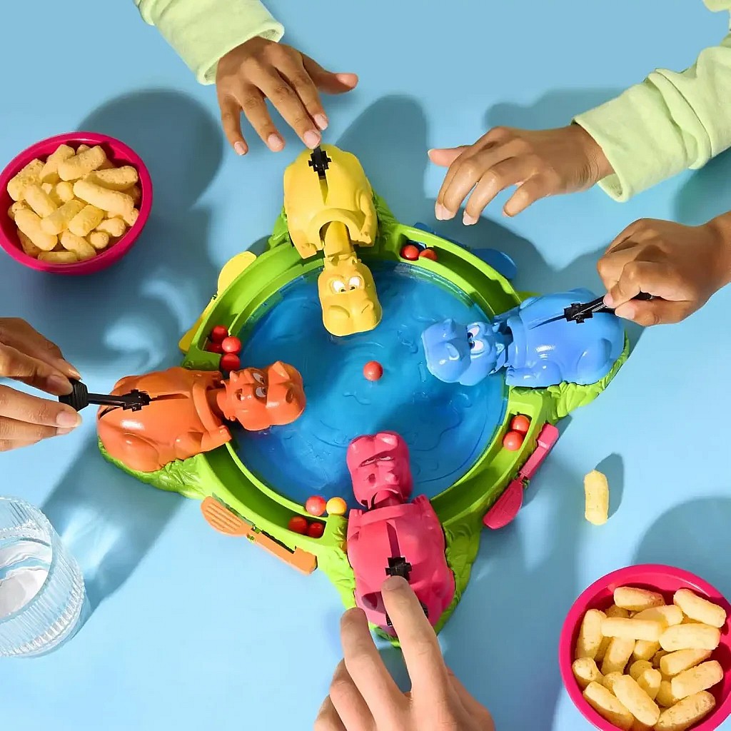 The images show two contrasting board games. The first is a vibrant, overhead shot of four players' hands pressing the levers of the yellow, blue, orange, and pink hippos in the classic children's game Hungry Hungry Hippos, with red marbles in the center and two bowls of puffed snacks nearby on a blue surface. The second is the cover of the Aeon's End (Second Edition) board game box, featuring a group of mages battling a massive, red, horned demon.