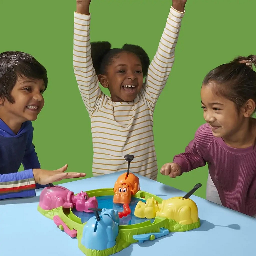 three excited children playing the Hungry Hungry Hippos game on a blue table against a green background, with the girl in the center cheering with her hands raised as her orange hippo appears to have won. The second image is the box cover for the cooperative deck-building game Aeon's End (Second Edition), featuring a group of mages battling a massive, red, horned demon.