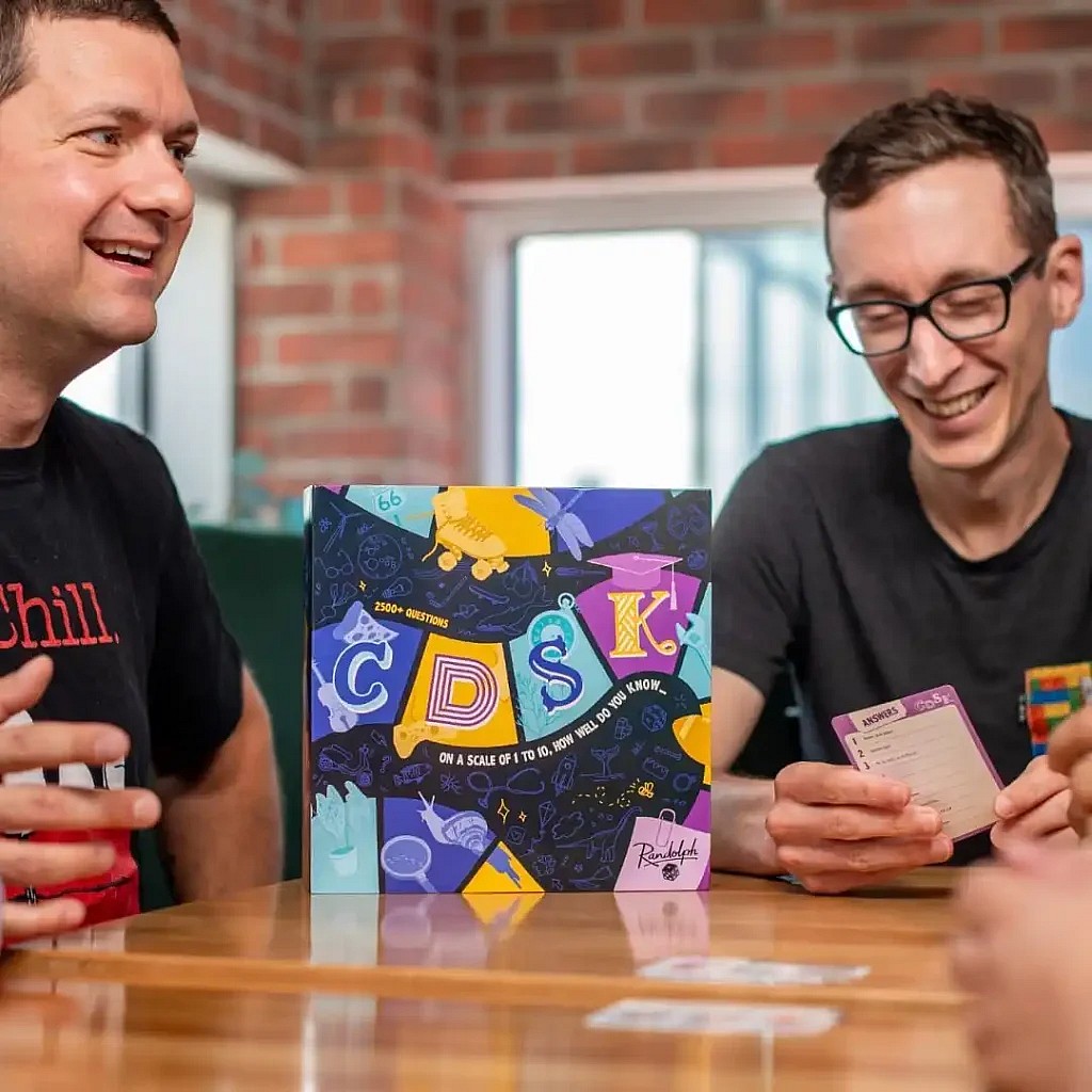 Two laughing people sit at a table playing a trivia game, with the brightly colored Trivial Pursuit: Decades box centered between them, and one player holding an answer card.
