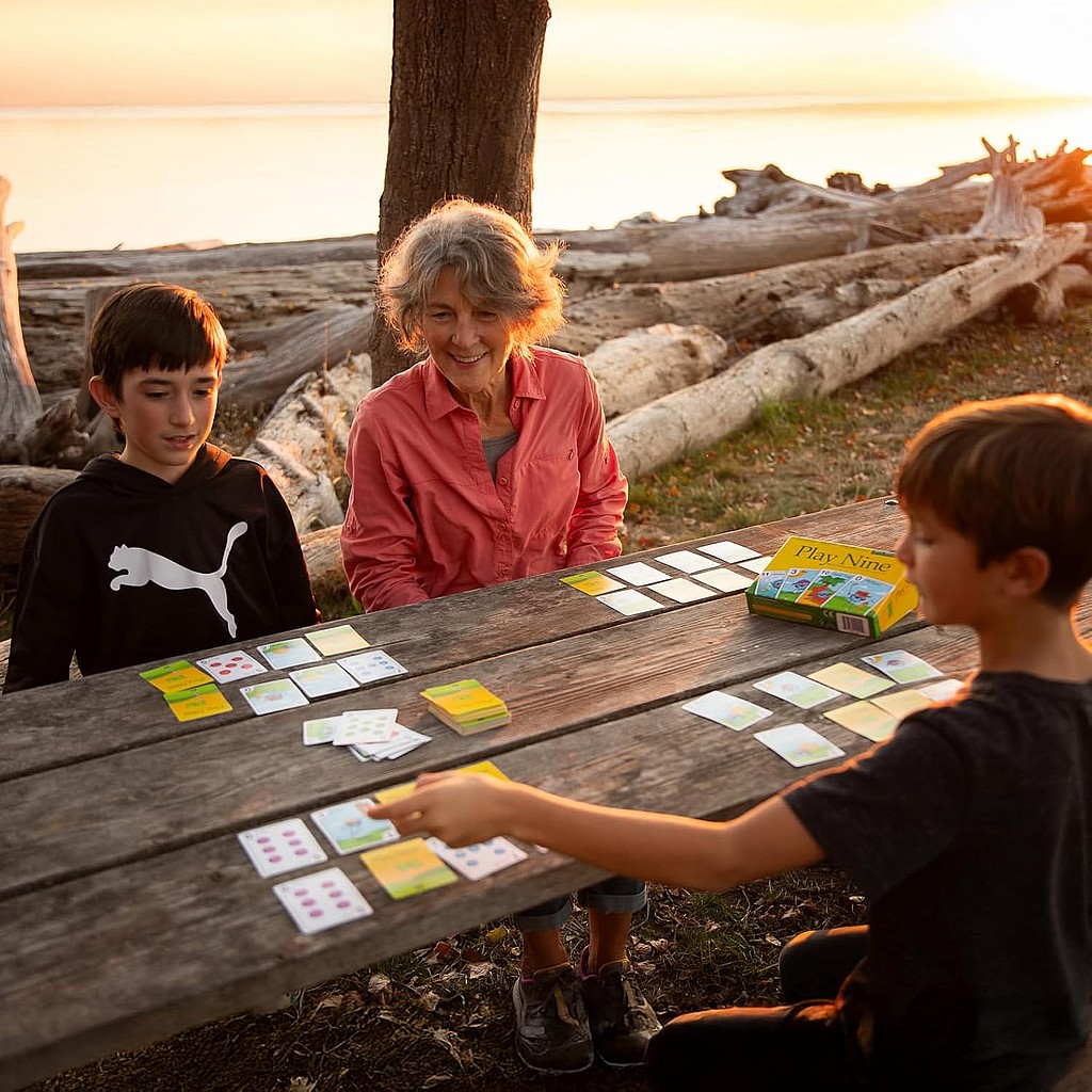 a grandma and her two grandsons play the card game "Play Nine: The Card Game of Golf" in a picnic wooden bench outdoors in the sunset