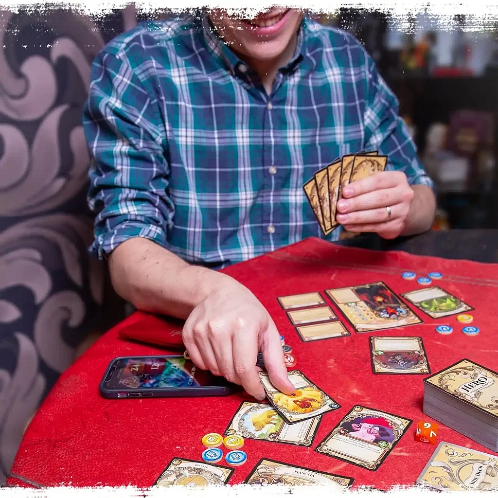close up of a person playing a card in the game Hero: Tales Of The Tomes card game on a red cloth table with the whole game setup