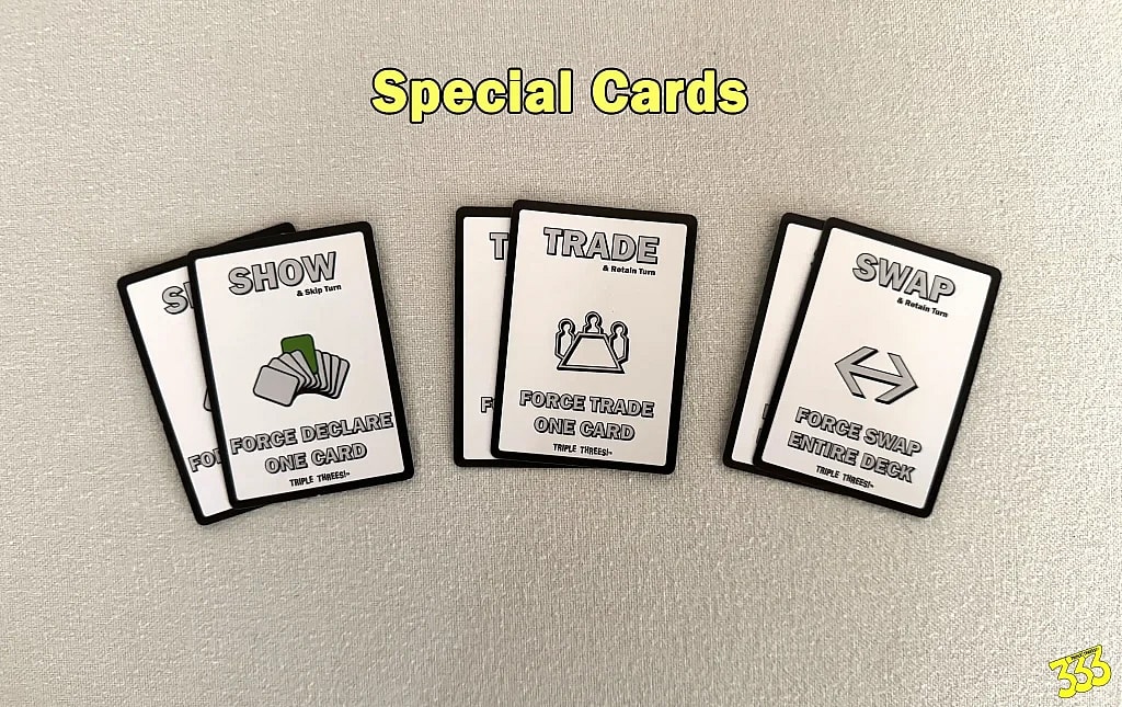 A flat-lay photo showing three types of special cards from the game "Triple Threes!". Each card is white with a black border and simple icons. The title "Special Cards" is at the top of the image.
The stack on the left is the "SHOW" card, with a symbol of a hand of cards. The text reads, "Force Declare One Card."
The middle stack is the "TRADE" card, featuring two hands shaking. The text says, "Force Trade One Card."
The stack on the right is the "SWAP" card, with a large horizontal arrow pointing in both directions. The text says, "Force Swap Entire Deck."