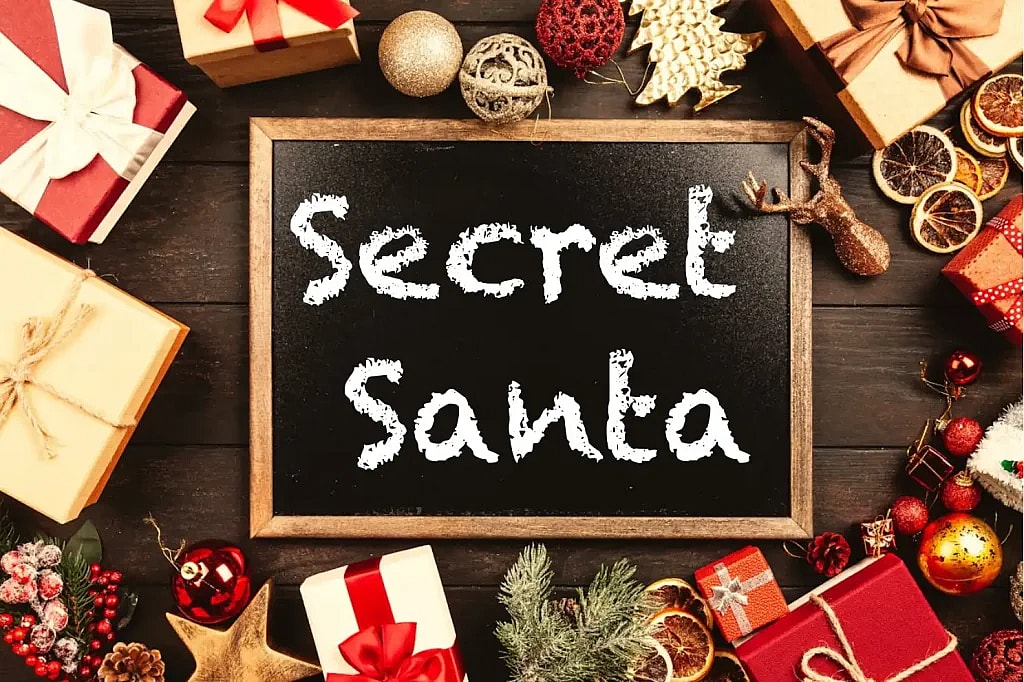 A flat-lay photograph of a small, rectangular chalkboard centered on a dark wooden surface, surrounded by festive Christmas decorations and wrapped gifts. The chalkboard has the words 'Secret Santa' written on it in white, chalk-style font. The surrounding decorations include ornaments (gold, red, and sparkly), dried orange slices, pine sprigs, a gold reindeer figurine, small wrapped presents with red and gold bows, and a gold star.
