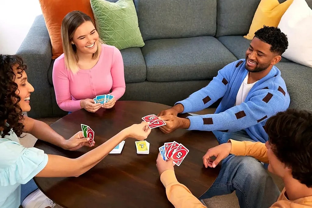 A diverse group of four young adults is sitting around a round coffee table in a living room, smiling and laughing as they play the Uno card game. Two people on the left side of the table are on one team, and the two on the right side are on the other. They are all holding cards and interacting with each other. A woman with a pink sweater is looking at a man in a blue cardigan and laughing as he passes her a card.