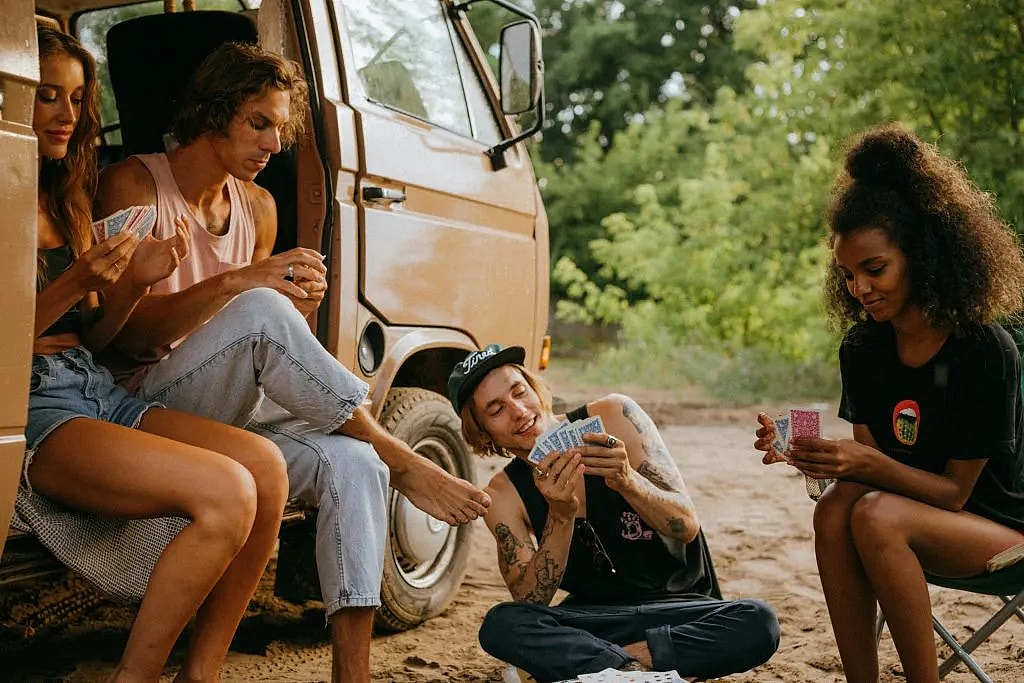 Young campers playing card games (bullshit or cheat) by a camper van in a forest setting, enjoying a summer adventure.