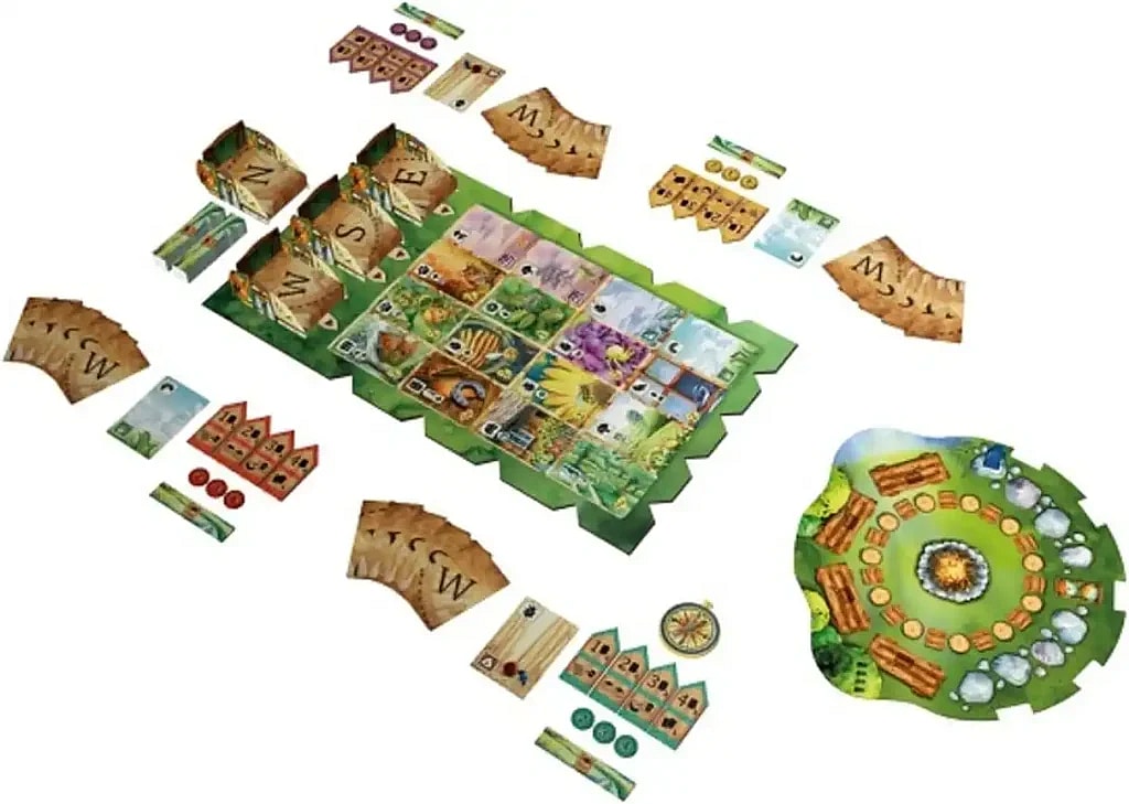 A bird's-eye view of the components laid out for the board game Meadow, set against a white background. The main central component is a grid of square cards depicting various natural elements like flora and fauna, which sits on a larger, irregular green playing mat. Arranged around this central area are several other components, including four player boards that stand upright (labeled N, E, S, and W, suggesting a compass rose), stacks of curved, brown Discovery Cards (also marked with 'W' and 'M'), smaller decks of cards, wooden tokens (some red, some green, some shaped like tents or cabins), and a round, green cardboard piece that resembles a campfire area surrounded by logs and stones. The overall scene shows the full setup for gameplay.