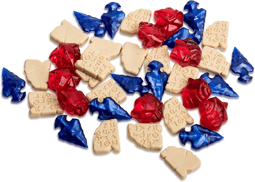The image displays a scattered pile of three types of plastic and cardboard resource tokens from the board game Lost Ruins of Arnak:
Red Translucent Jewels/Rubies: Clump-like, multifaceted translucent red plastic tokens.
Blue Translucent Arrowheads/Obsidian: Stylized arrowhead-shaped translucent blue plastic tokens.
Beige Tablets: Light beige, flat, irregularly shaped cardboard tokens printed with ancient-looking symbols or hieroglyphs. These tokens represent three of the higher-value resources used for advancing on the research track and defeating guardians in the game.