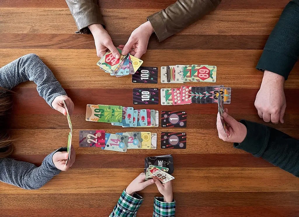 An overhead shot shows four people playing "The Game" card game on a wooden table. A pair of adult hands at the top of the frame holds a fan of cards. A child's hands at the bottom of the frame hold a small stack of cards. Two other players, one on the left and one on the right, are visible by their arms and hands, each holding a few cards.
Cards are laid out in four rows on the table between the players. Each row has a card with a small arrow on it, pointing up or down. A few cards are stacked on top of each other in the two middle rows. In the top row, a pile of cards in a hand is visible, with the top card showing the number 84. A card in the second row is numbered 100. A card in the third row is numbered 1. A card in the fourth row is numbered 22.