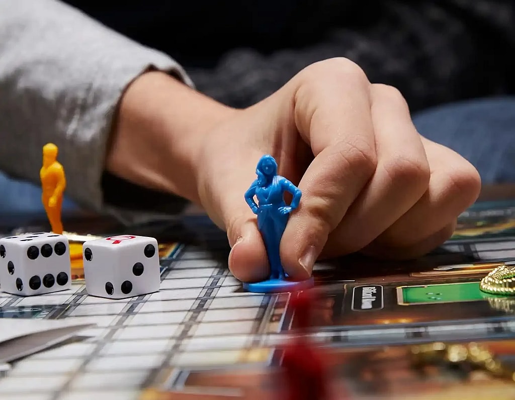 Close up of a hand moving a murder suspect token from the board game clue. game board, dice, and cards visible in the foreground