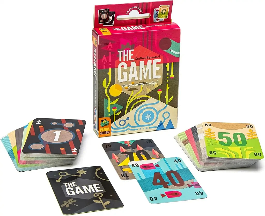 An angled, close-up shot of the card game "The Game" by Pandasaurus Games, set against a white background. The game's vibrant red box stands upright in the back, featuring abstract, colorful illustrations and the text "THE GAME" in large, stylized font. Below the title, it reads "Are you ready to play?".
In the foreground, the game's components are laid out. To the left is a thick stack of cards with a single card on top displaying a large "1" and several circles. To the right is another stack of cards with the top card showing a large "50". Between the two stacks, a few cards are fanned out, one showing a large "40" with fish icons and another showing a large "70" with abstract plant or flame-like icons. A single black card with a floral design and the title "THE GAME" sits in the center foreground.