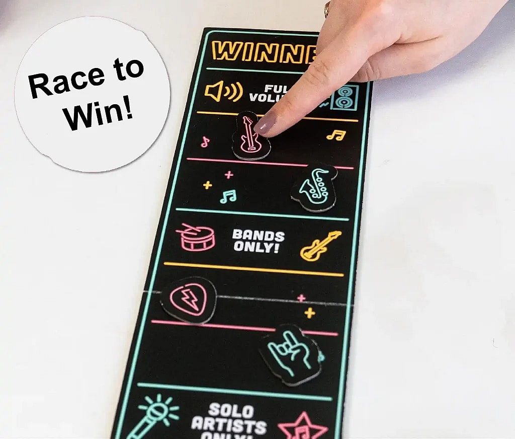 An overhead shot of Grab the Mic game board. Shows a person's hand with painted nails pointing to a magnetic piece shaped like an electric guitar on a black game board. The game board is decorated with pink, yellow, and blue neon-style outlines of various musical items and has the word "WINNER" at the top. Magnetic pieces are scattered along the board, including a drum set, a microphone, a saxophone, and a hand making a "rock on" gesture. A white circle with the words "Race to Win!" is superimposed on the top left of the image.