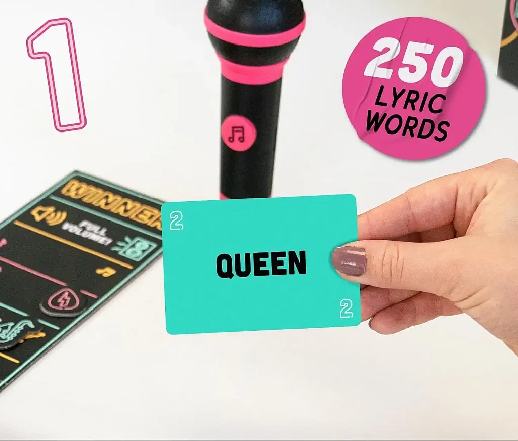 An image shot from a low angle, showing a hand holding a turquoise card with the word "QUEEN" in bold black letters. The card has a small number 2 in the top left and bottom right corners. In the background, there's a black and pink toy microphone. To the left, a game board or player mat is visible, marked with the words "WINNER" and "FULL VOLUME." The number 1, a round icon that reads "250 LYRIC WORDS" in white text on a pink background. Grab the Mic party card game