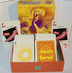 A photograph of the card game Canasta Caliente shows the box and some of the cards. The tall, yellow box features a silhouette of a dancing figure in a swirling purple and yellow background. Text on the box includes "Canasta Caliente OFFICIAL VERSION" and "South America here we come! Play a little CARDS! Have a lot of FUN!" In front of the box, two face-up cards are visible, both with a value of "-100" and an illustration of a red chili pepper on a black background, labeled "CALIENTE." Two stacks of face-down cards are sitting in an orange tray or half-box. The top card of the left stack is an Ace card with a value of "20" and a large yellow sun illustration. The top card of the right stack shows the intricate orange and red patterned back of the cards, which includes the words "Canasta Caliente." A green Hasbro logo is visible on the front of the orange tray.