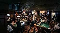 a dark, cinematic photo of a casino heist, with masked figures in tactical gear. One figure reaches over a craps table grabbing chips, while in the foreground, a broken security camera is smoking. Other armed, masked individuals are visible, including one pointing a firearm and another running toward a vault, creating a scene of tension and chaos.