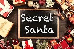 A flat-lay photograph of a small, rectangular chalkboard centered on a dark wooden surface, surrounded by festive Christmas decorations and wrapped gifts. The chalkboard has the words 'Secret Santa' written on it in white, chalk-style font. The surrounding decorations include ornaments (gold, red, and sparkly), dried orange slices, pine sprigs, a gold reindeer figurine, small wrapped presents with red and gold bows, and a gold star.