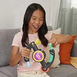 A happy young girl, with long dark hair, sits on a couch while playing with a Bop It! game. She is holding the black, yellow, green, and pink toy with both hands, positioned to pull the "pull it" handle on the left and twist the "twist it" knob on the right. She is wearing a light pink shirt and beige pants and has a surprised but excited look on her face. The couch behind her is gray with white and orange pillows.