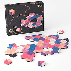 The image displays the board game "CUBED: Next Level Dominoes". The game box is visible in the background, which is black with a modern, wavy design. The front of the box has the game logo, and text that reads "NEXT LEVEL DOMINOES," "62 PIECES," "1-6 PLAYERS," and "AGES 5+." The game pieces are shown in two states: some are arranged on a white surface to form an abstract, colorful pattern, while others are scattered around the main formation. The pieces are three-dimensional, hexagonal prisms, with each side featuring a different color.