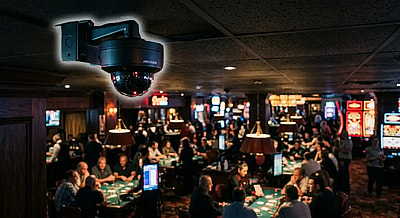 security camera outlined with a soft glow overlooking a casino floor with players and tables playing card games.