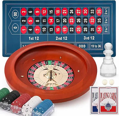 A high-angle shot shows a wooden roulette set with a felt betting mat featuring a double-zero American layout. The set includes a professional-style wheel, stacks of colorful poker chips in a clear tray, two decks of playing cards, two white roulette balls, and a clear plastic win marker.