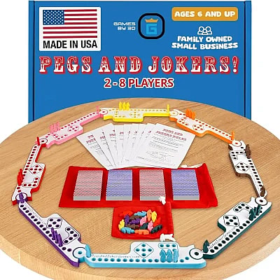 An image of the Pegs and Jokers board game set displayed on a wooden table, featuring colorful interlocking plastic game tracks arranged in a circle for 2-8 players. The set includes four decks of playing cards, several rule sheets, and a variety of colored horse-shaped game pieces. The background shows the blue game box, which highlights that the product is Made in USA by a family-owned small business and is intended for ages 6 and up.