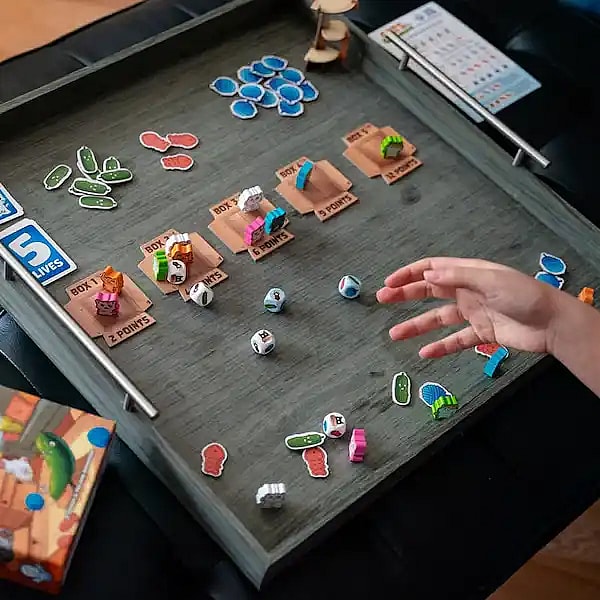 a person's hand reaching toward a tabletop board game, Cats vs Cucumbers, which is set up inside a large grey wooden tray. The game components include colorful cat meeples placed on small cardboard boxes labeled with point values, scattered cucumber and yarn tokens, multiple custom dice, and scoring cards.