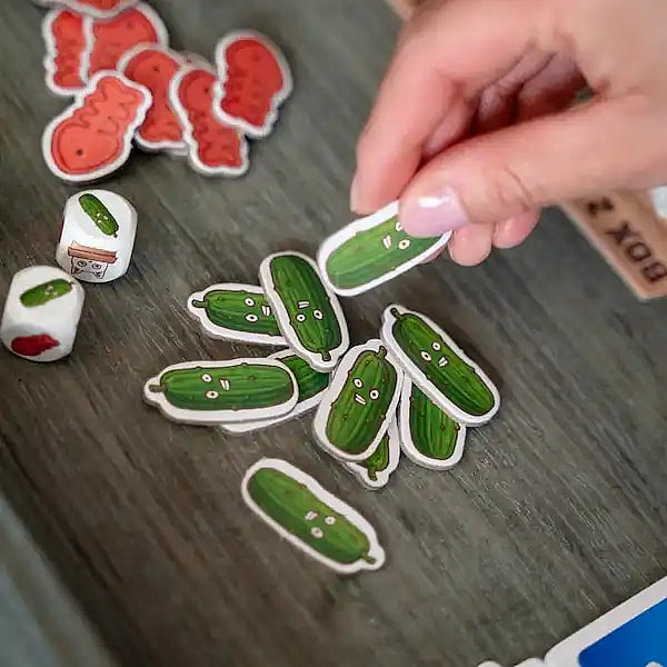 hand holding a cucumber token from the board game Cats vs Cucumbers. Dice, and other tokens are visible in the background