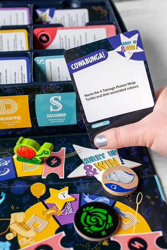 A close-up of the Trivial Pursuit: Decades game, showing a hand holding a question card that reads "COWABUNGA! Name the 4 Teenage Mutant Ninja Turtles and their associated colours." In the foreground are game pieces, including a green turtle token, and category dividers (D, S) in the game box.