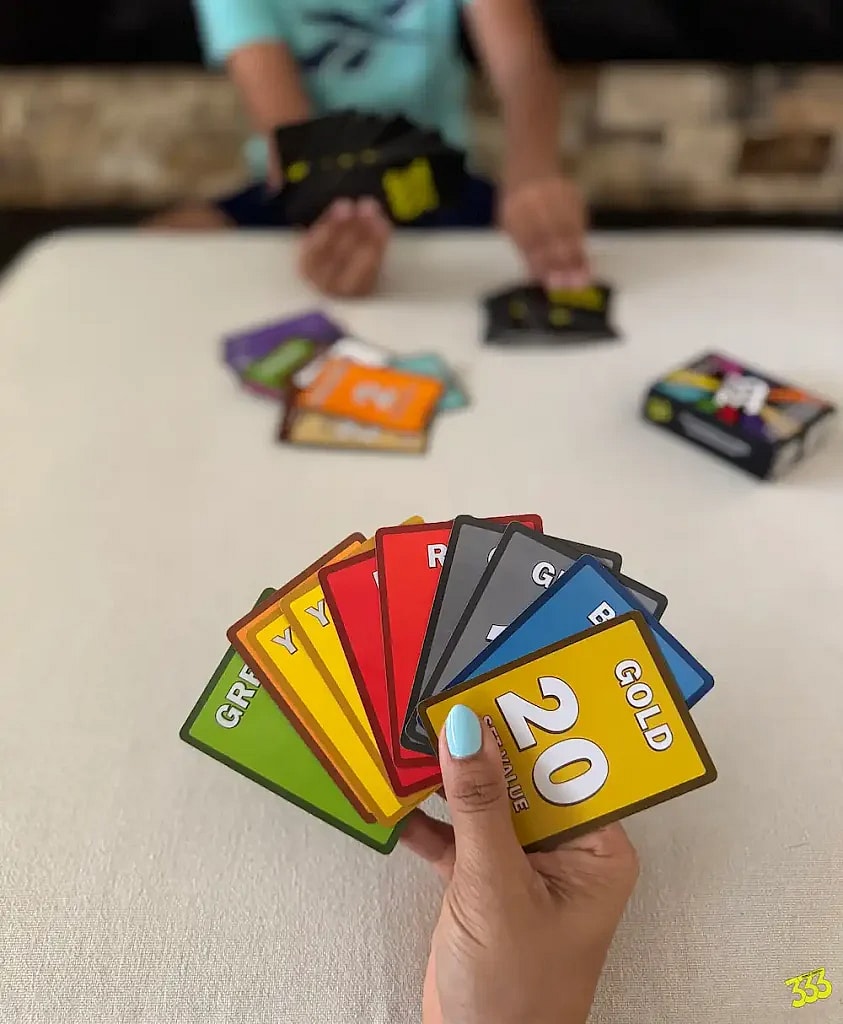 A first-person perspective shot of a person's hand holding a fan of playing cards from the card game "Triple Trees!". The cards are from a game called "Triple Threes!" and are different colors, including green, yellow, red, gray, blue, and a special gold card with the number 20 on it. The player's fingernails are painted light blue. In the background, another person, possibly a child, is also holding cards and sitting across a table with other cards scattered between them. A game box is visible in the distance.