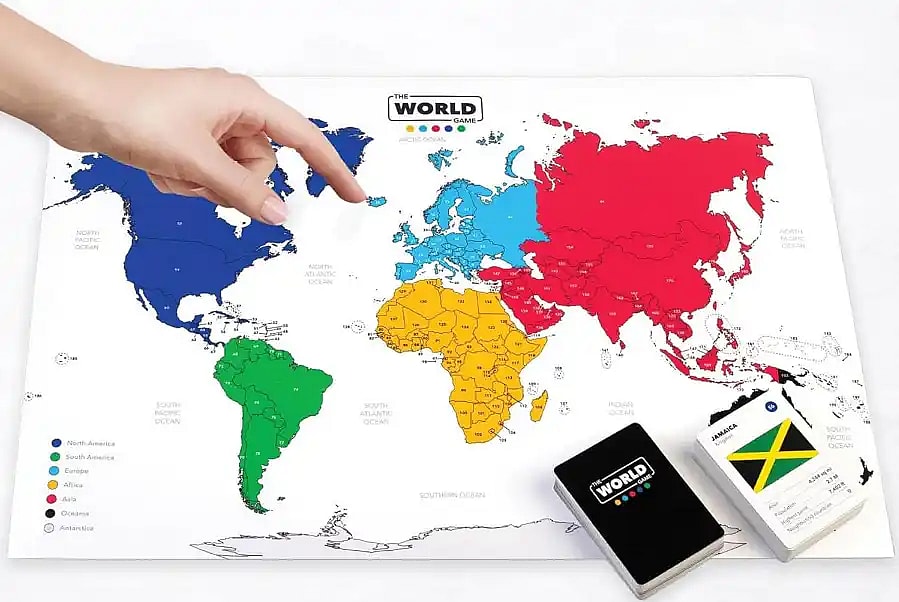 A hand pointing at a colorful world map board from The World Game geography board game, shown alongside a deck of trivia cards featuring a card for Jamaica.